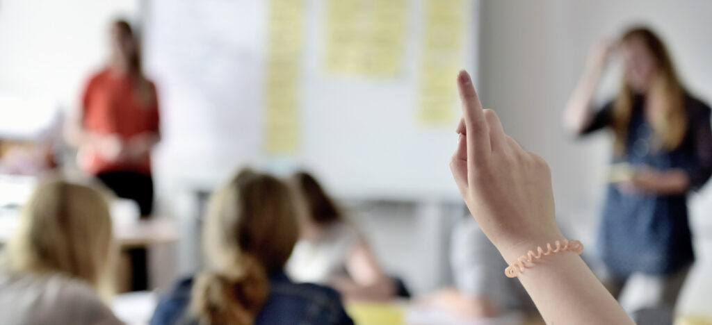 Immagine di una mano che alza la mano durante una lezione in classe con studenti seduti, scrivanie e lavagna sullo sfondo