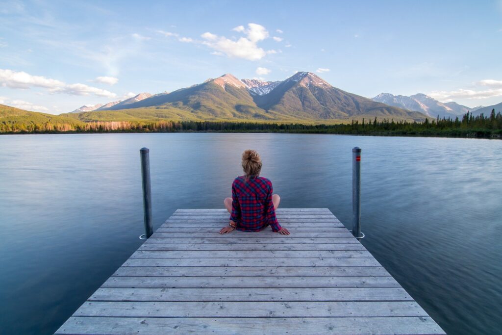 Donna seduta su un pontile di legno che guarda il湖 con montagne e paesaggi naturali mozzafiato sullo sfondo, creando un'atmosfera di serenità e tranquillità